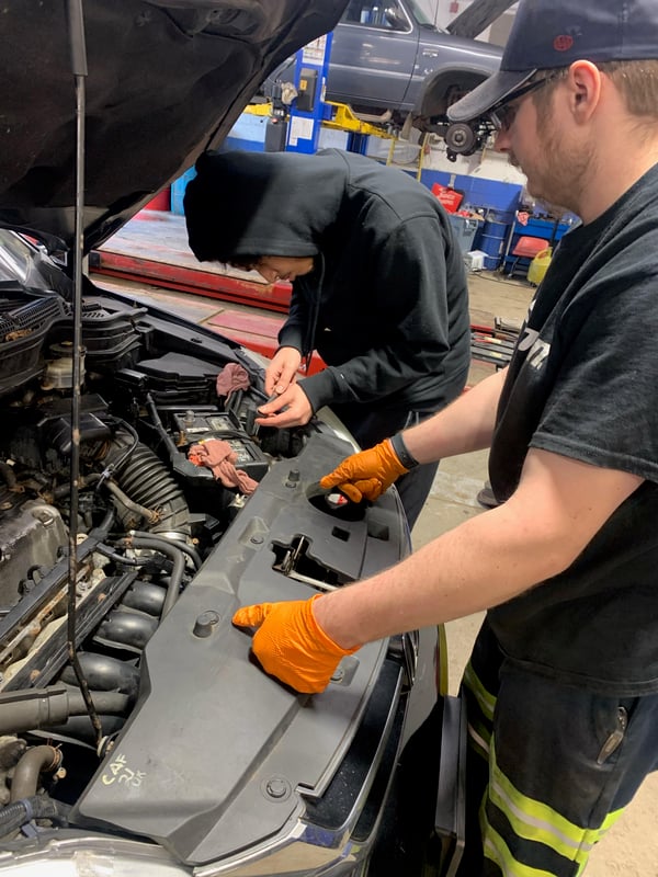 A student changing a car battery being supervised by his mentor.